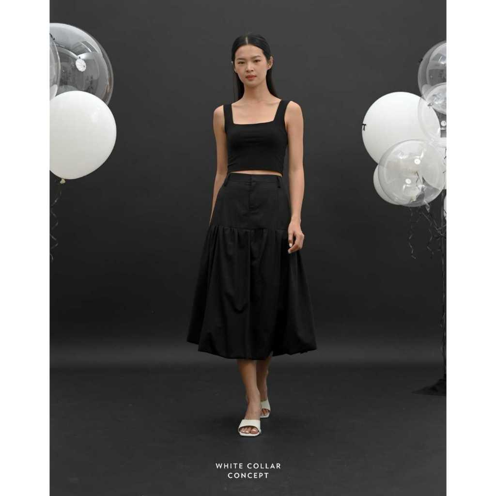 A woman standing in a studio setting, wearing a black tank top and a black bubble skirt with a short waist, next to balloons and a reflective sphere.