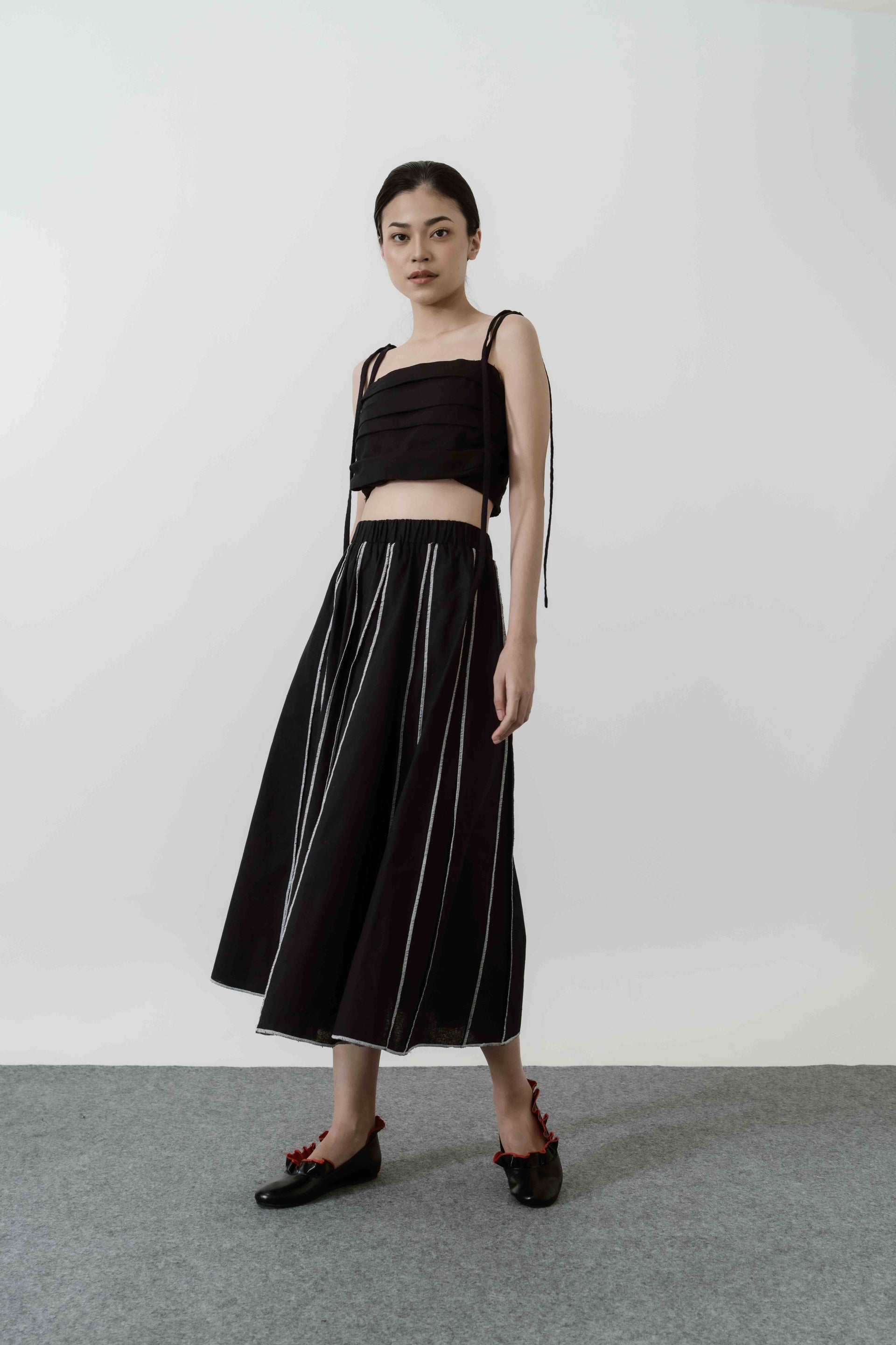 A woman standing wearing a black midi skirt by Morningsol with a flared design, paired with a black top and black shoes.