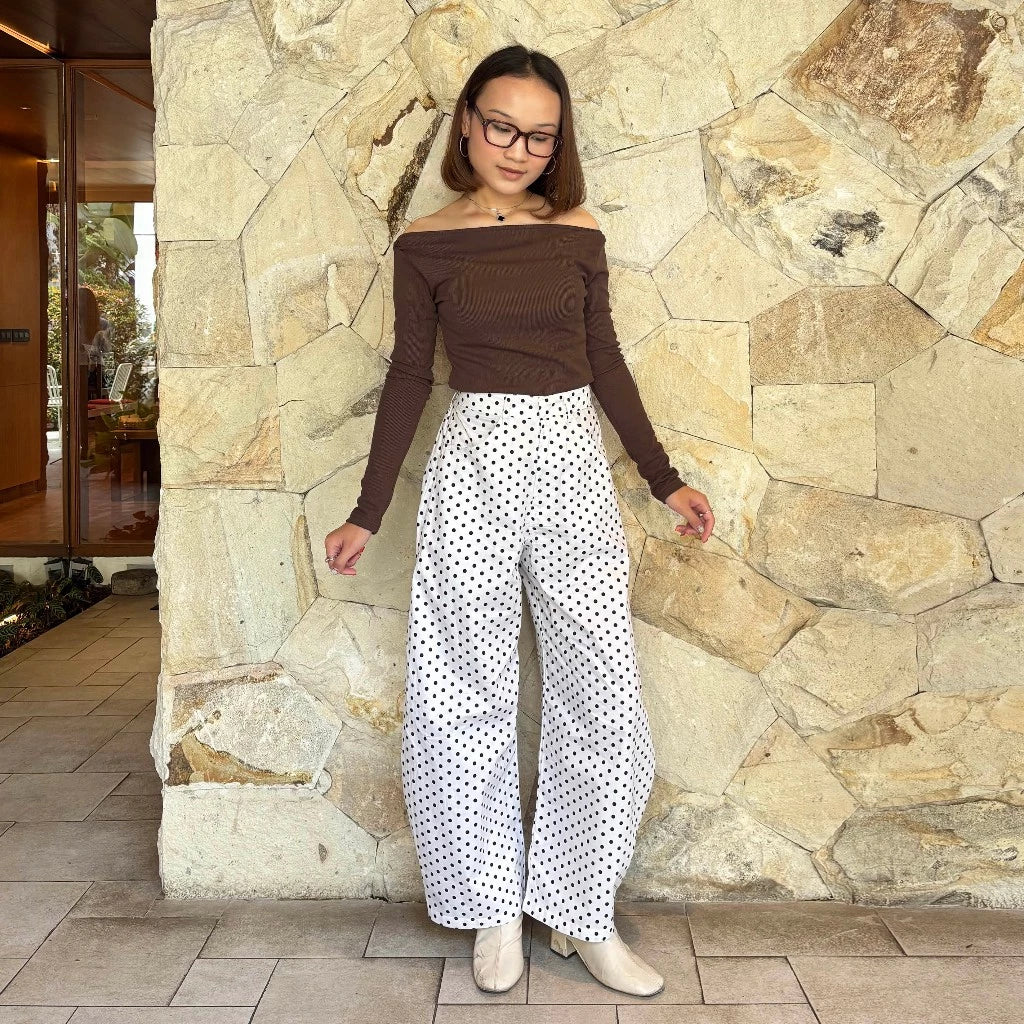 Woman wearing a brown top and white polka dot barrel pants standing against a stone wall.