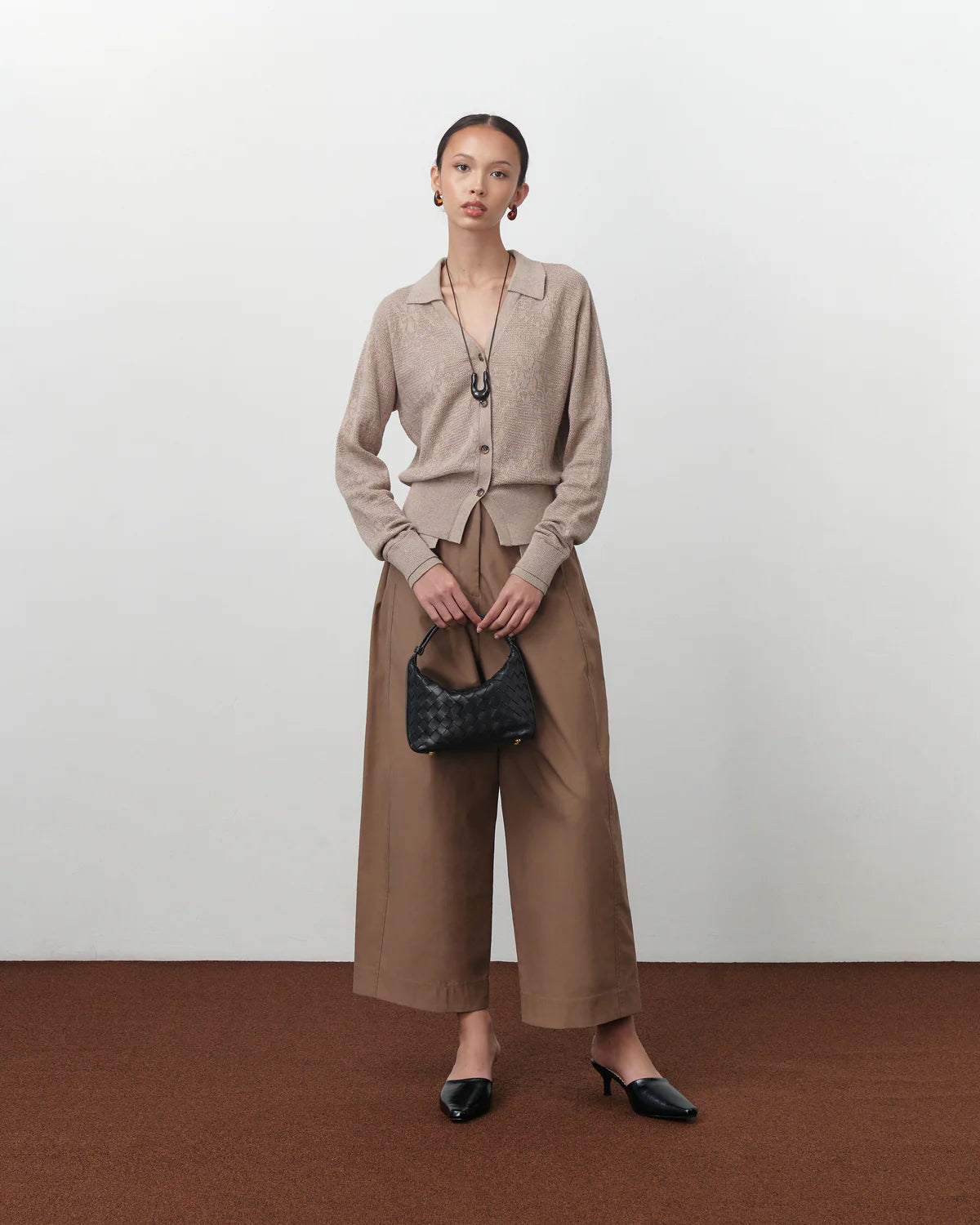 Woman wearing a brown cardigan by Achete De Nous with wide-leg pants holding a black handbag against a white background