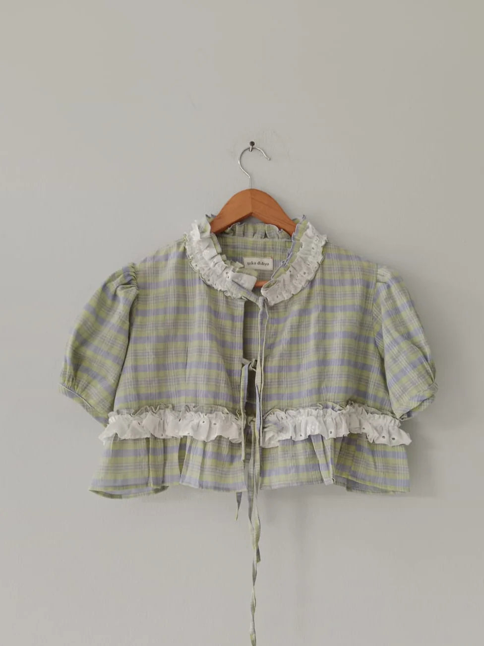 Plaid jacket by Toko Didiyo with ruffled details on a hanger against a plain background