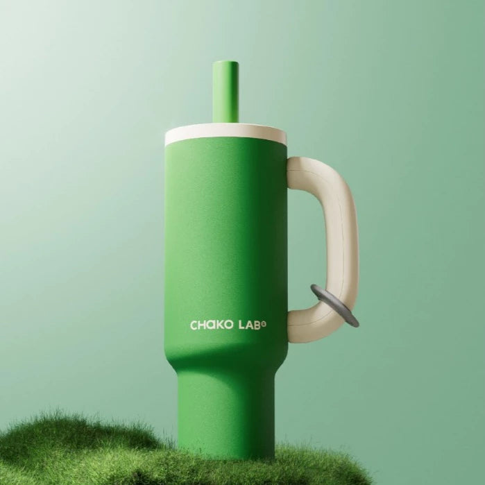 A green Chako Lab mug with a white handle and green straw, placed on a rock with flowers in the background.