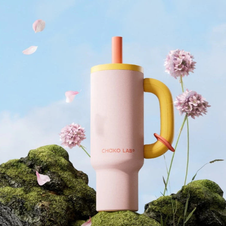 A peach Chako Lab tumbler with a yellow handle, placed on a rock with flowers in the background.