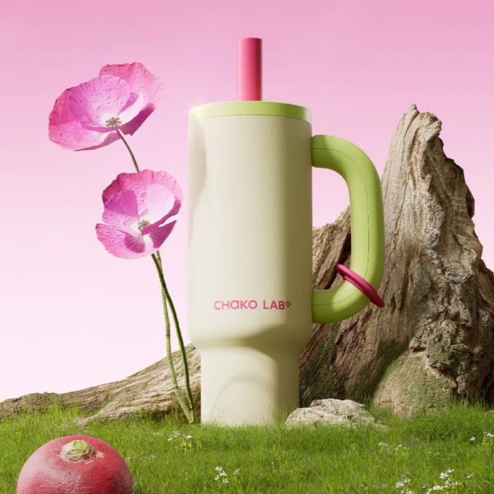 A light green Chako Lab tumbler with a green handle and pink straw, placed on a rock with flowers in the background.