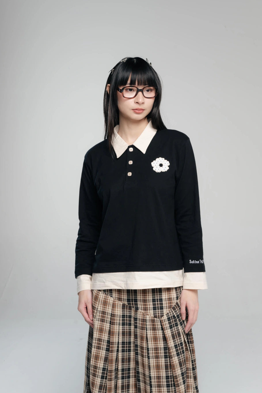 A model wearing a black long-sleeve polo by Tome Ame with a cream collar, cream hem, and a white flower applique on the chest, paired with a brown and beige plaid skirt, against a solid gray background.
