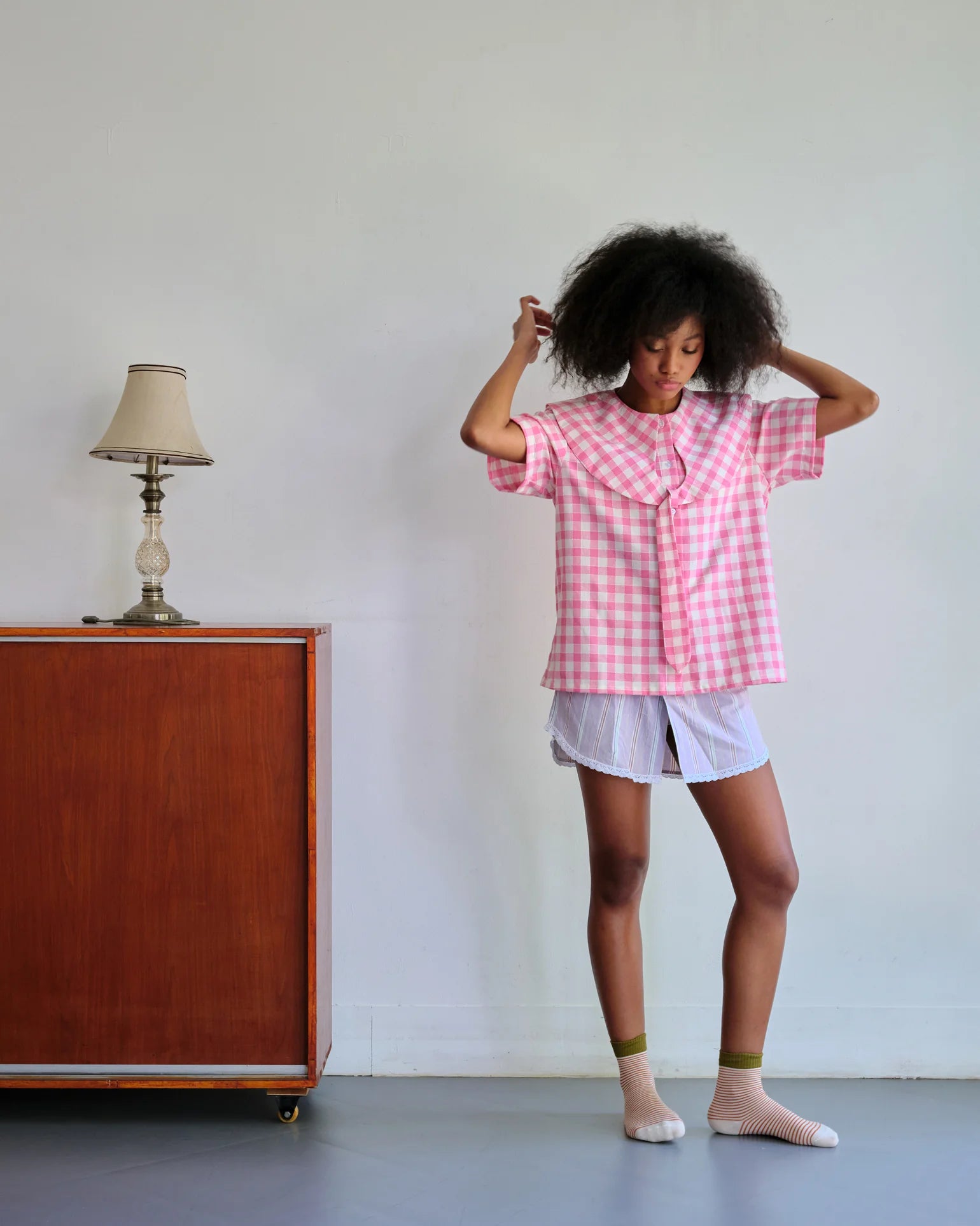 Woman wearing a pink checkered shirt by Toko Didiyo and white shorts standing in a room with a wooden cabinet and lamp.