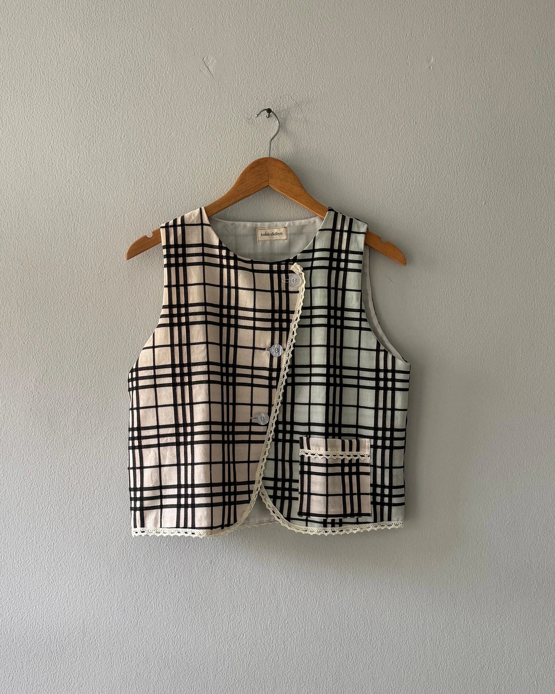 Black and white checkered vest from Toko Didiyo made from linen tartan pattern and lace trim details along the hem on a wooden hanger against a plain wall.