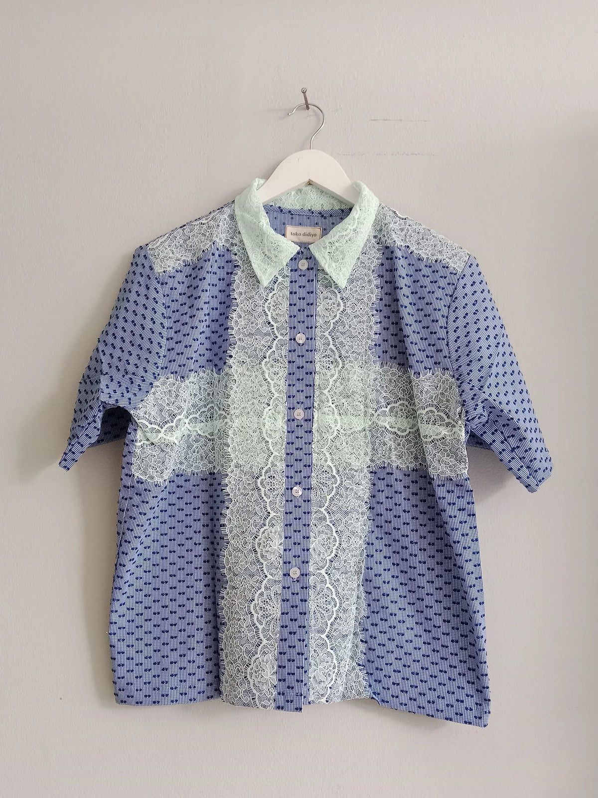 A short-sleeve button-up shirt by Toko DIdiyo hanging on a white hanger against a plain light gray wall. The shirt features a light blue base with a small dark blue geometric pattern, accented by white lace panels running vertically down the front and across the shoulders.