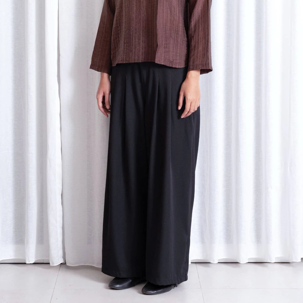 Person wearing a brown top and black wide-leg pants notyourcompany standing against a white curtain.