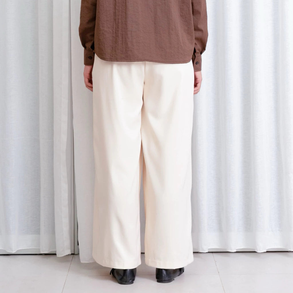 Person wearing a brown top and cream wide-leg pants by notyourcompany standing in front of white curtains.