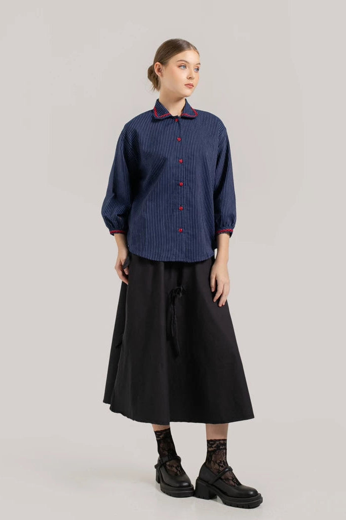A person standing against a plain background, wearing a dark blue denim striped shirt with a collared neckline and buttons down the front by Tuff Puff, paired with a black skirt and black ankle boots.