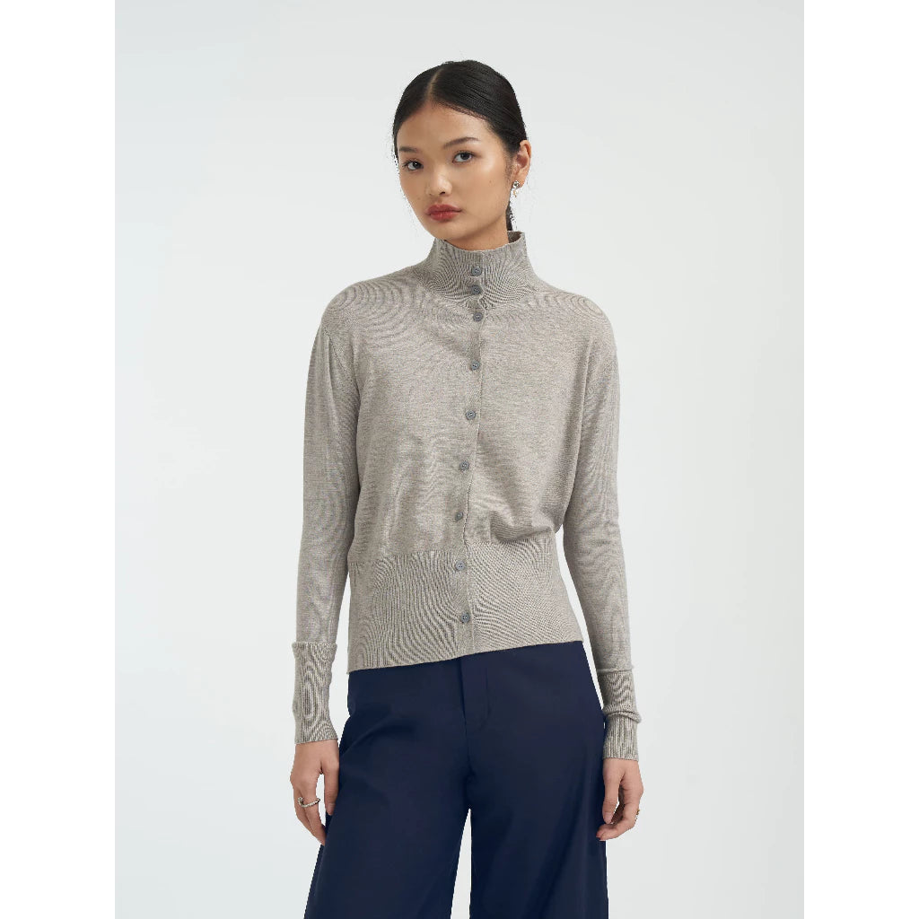 Woman wearing a patterned grey turtleneck cardigan by Achete de Nous and dark pants on a white background
