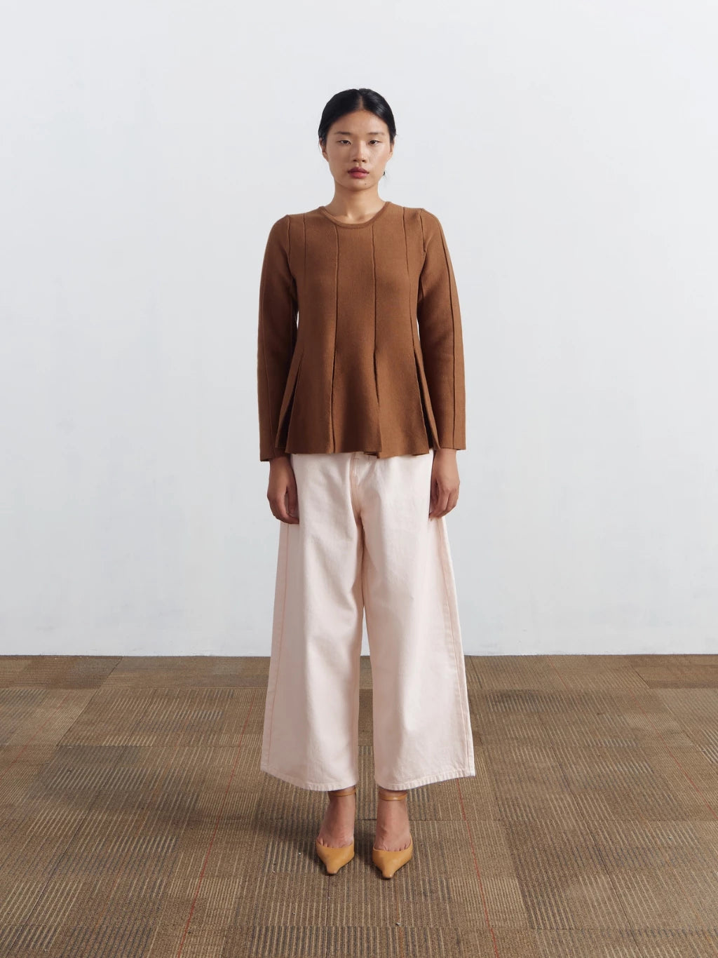 Woman wearing a brown longsleeved blouse by Achete de Nous and beige pants standing on a wooden floor with a white background