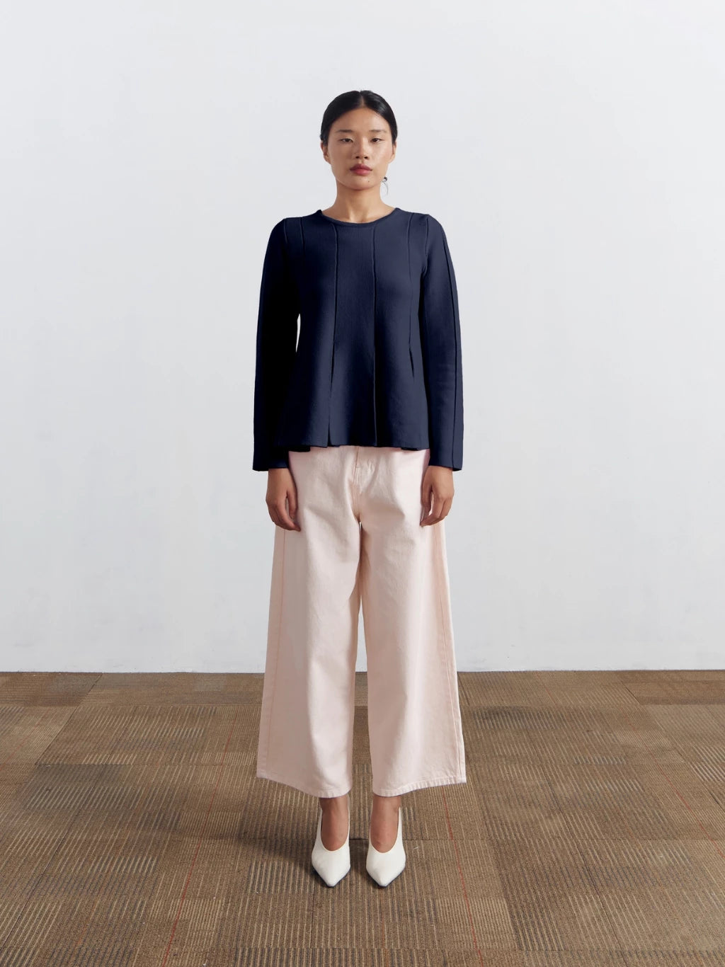 Woman wearing a navy blue longsleeved blouse by Achete de Nous and beige pants standing on a wooden floor with a white wall background