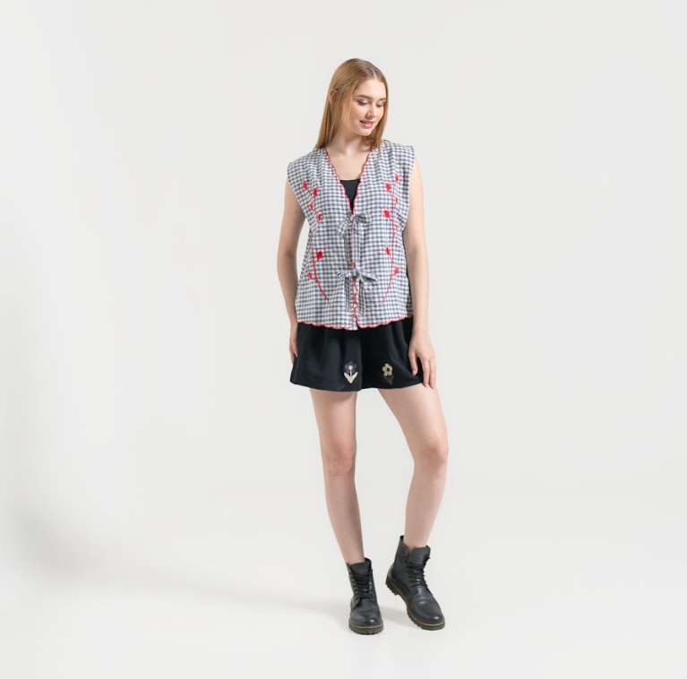 Woman wearing a checkered vest with red flower embroidery over a black top and shorts on a white background