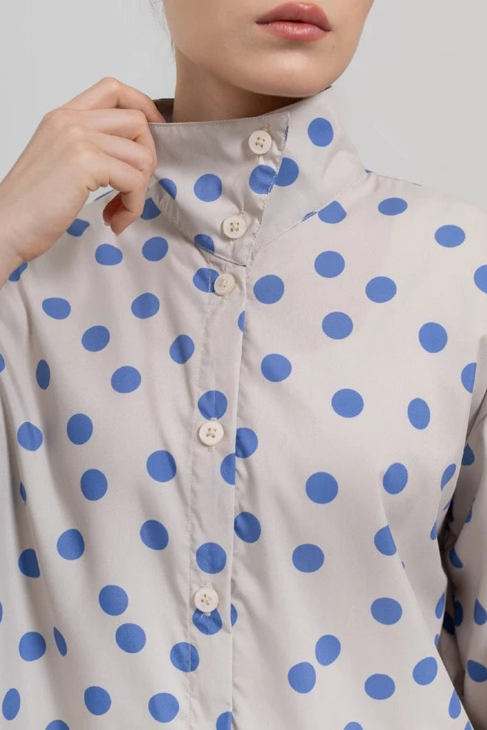 A close-up of a person wearing a light beige or off-white long-sleeve button-front shirt with large blue polka dots and a high stand collar by Tuff Puff. The person's hand is adjusting the collar, and the background is a plain light gray studio backdrop.