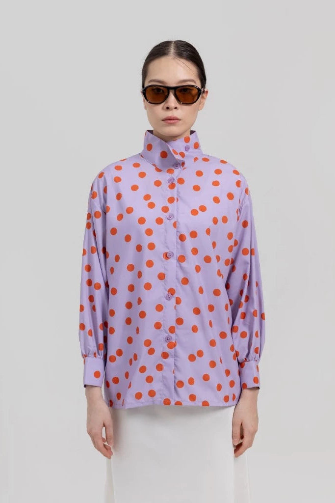A model wearing a lavender long-sleeve button-up shirt with orange polka dots pattern by Tuff Puff, paired with white trousers and dark sunglasses, against a solid light gray background.