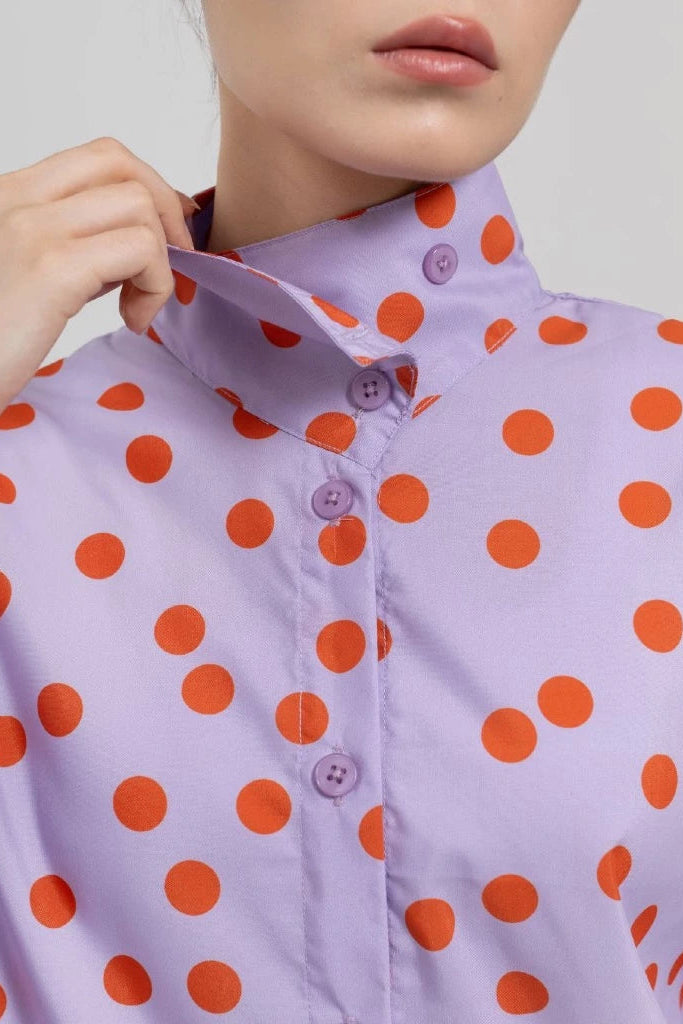 A close-up of a model wearing a lavender button-up shirt with a bold orange polka dots pattern by Tuff Puff. The model is adjusting the collar, showing the fabric texture and the buttons at the neck.