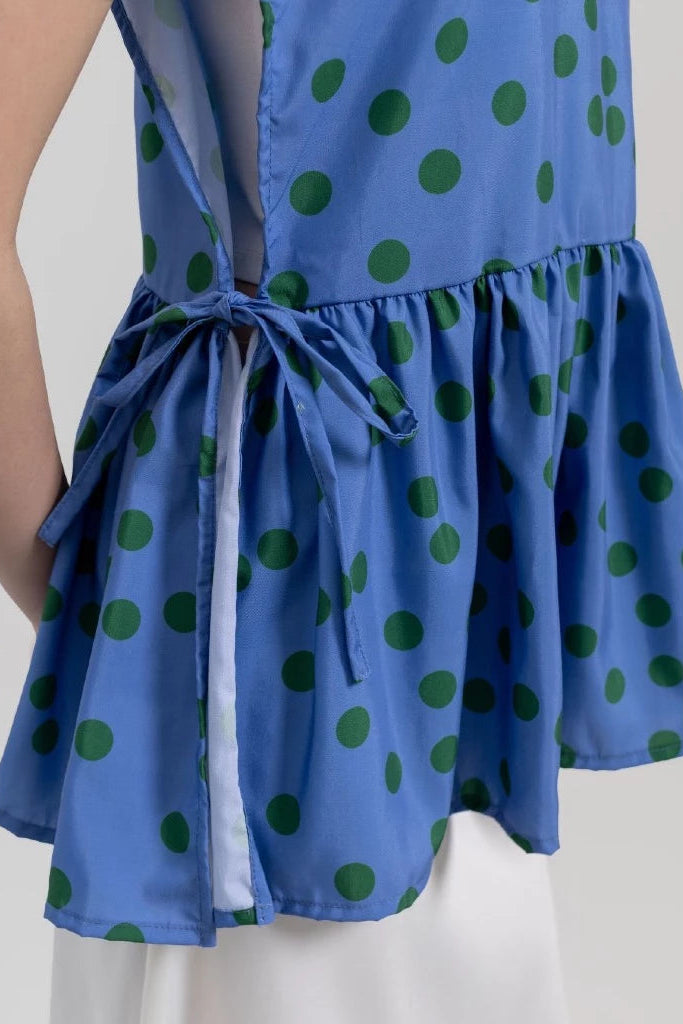 A close-up of a sleeveless blue vest with green polka dots by Tuff Puff, featuring a tie-waist detail and a ruffled peplum hem, worn by a model.