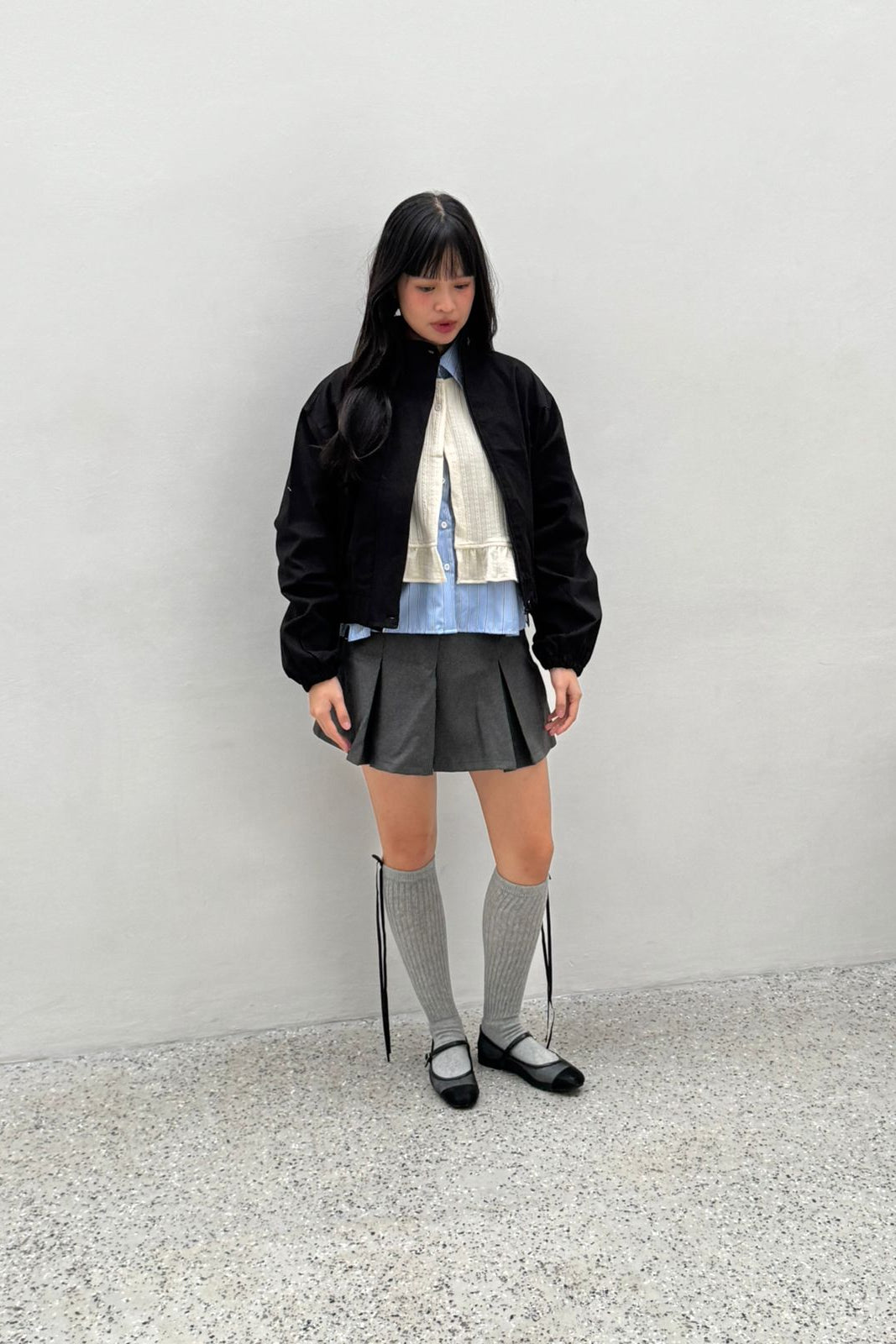 Person wearing a black jacket by notyourcompany, patterned shirt, and gray skirt against a white wall.
