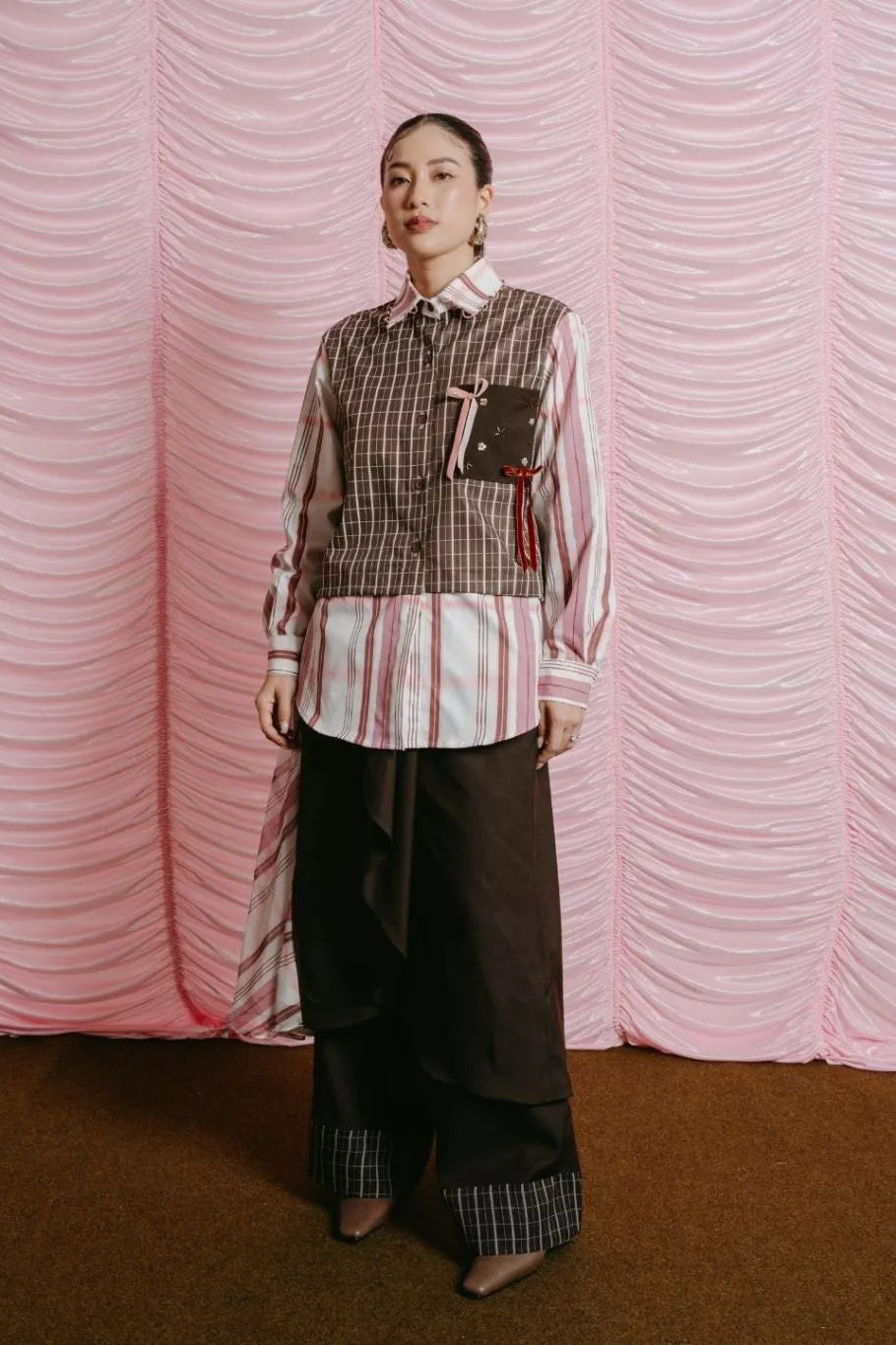 Person wearing a checkered vest by LR Studio over a striped shirt with wide-leg pants against a pink textured curtain.