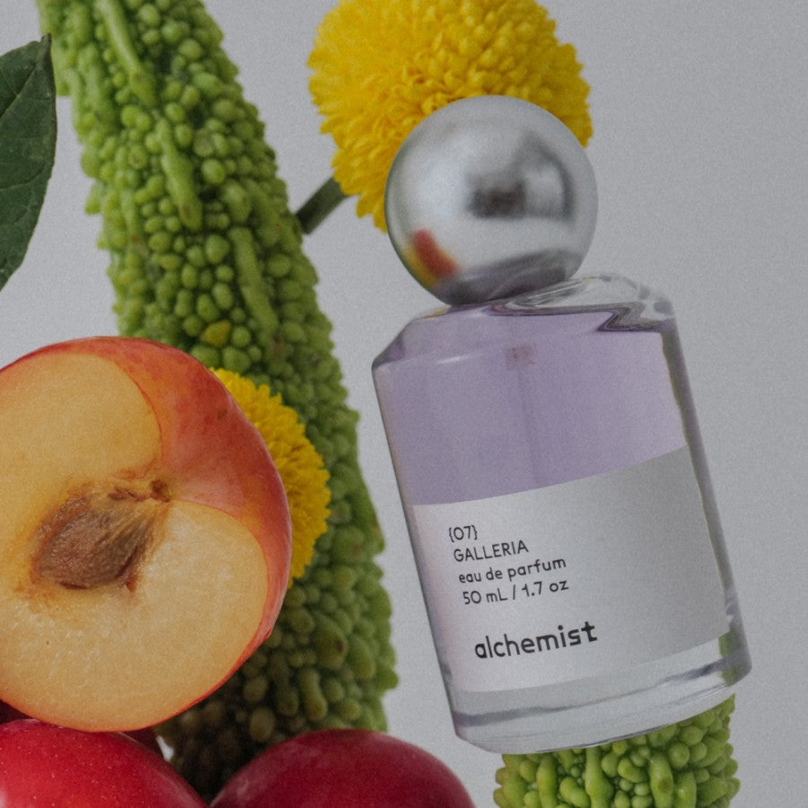 A bottle of 'Galleria' fragrance by Alchemist placed among floral and fruit elements, suggesting a connection to the natural world and the ingredients of the perfume.