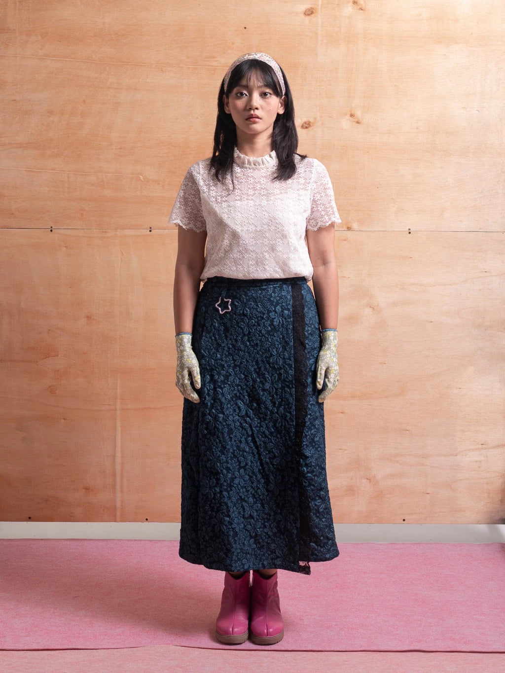 Woman wearing a white lace top, dark blue patterned skirt, and pink shoes against a wooden wall.