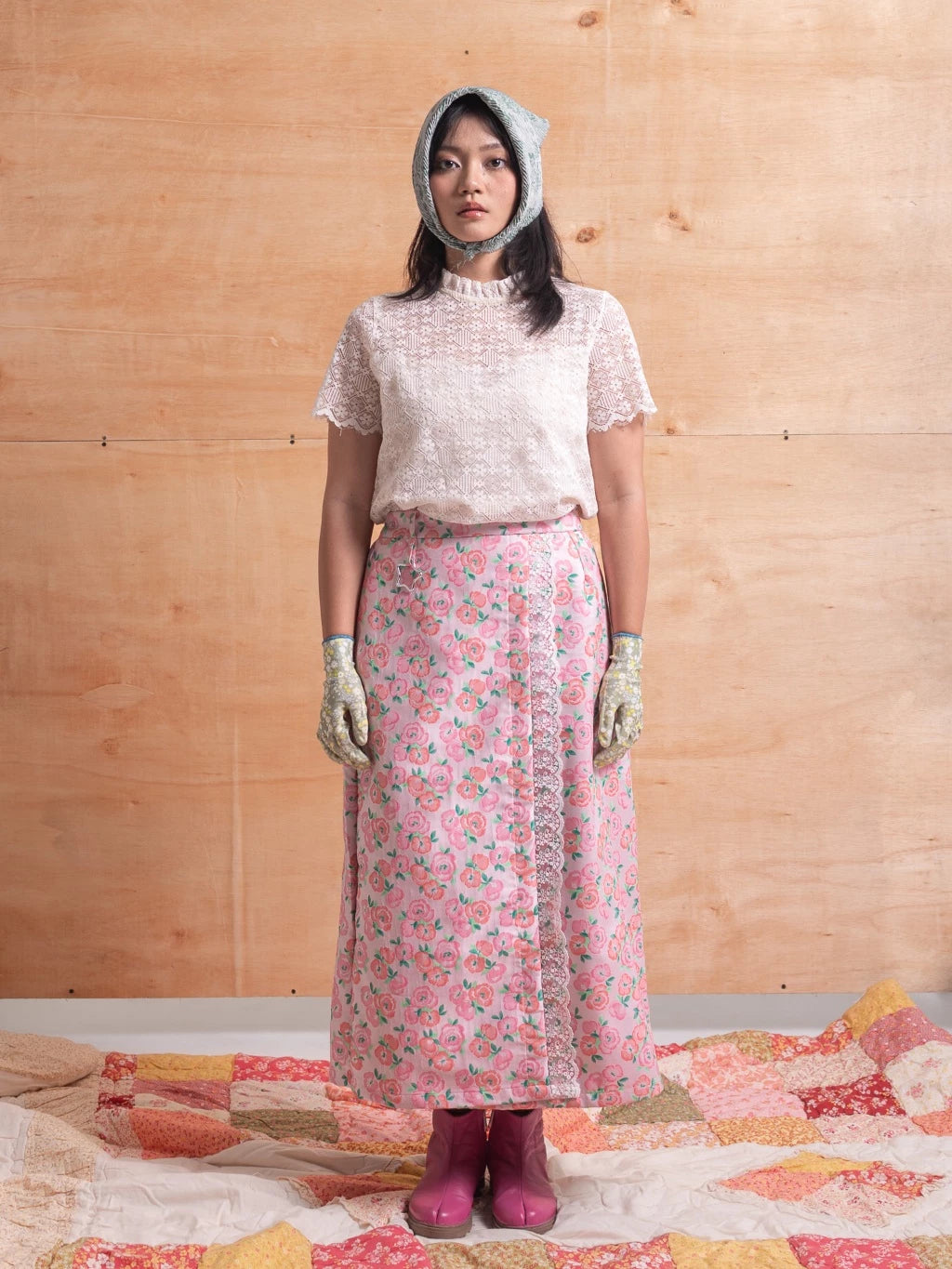 Woman wearing a pink floral skirt, white top, and pink boots standing on a quilted surface with a wooden wall background.