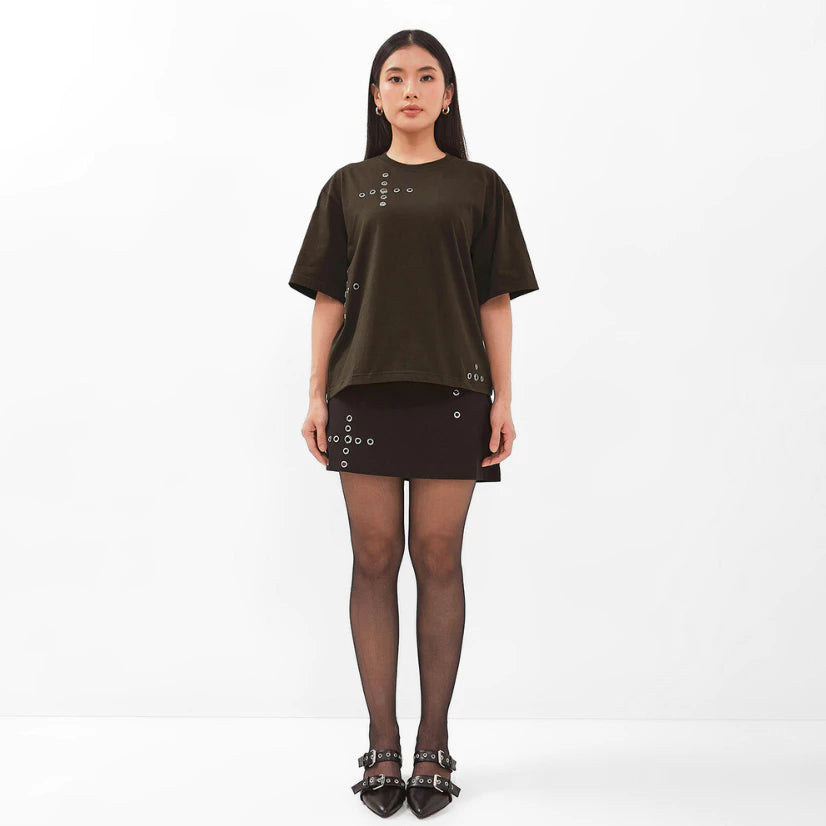 Woman wearing a brown oversized t-shirt by Isoos Studios and black skirt with studded details on a white background