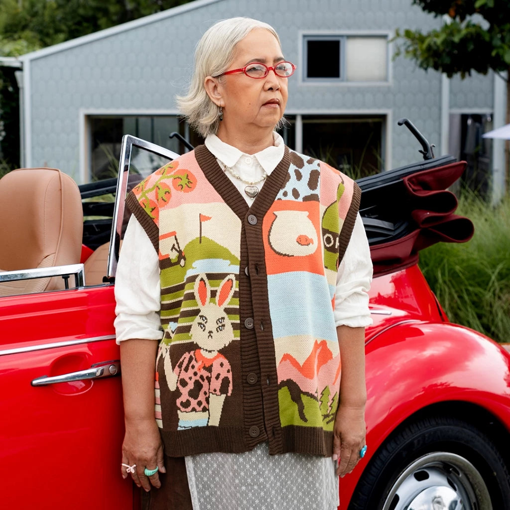 Woman wearing "Golf Club" Kintted Vest, a knitted vest with colorful graphic design featuring a golf course, bunny illustration, and playful patterns from Oh.Irv, worn over a white collared shirt by an older woman standing beside a red convertible car in an outdoor setting.