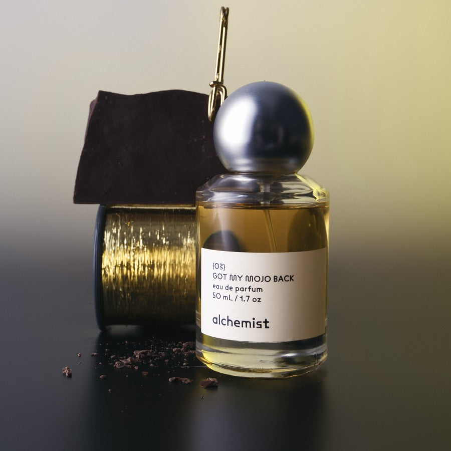 A bottle of 'Got My Mojo Back' fragrance by Alchemist with a metallic cap, accompanied by a gold-colored object.