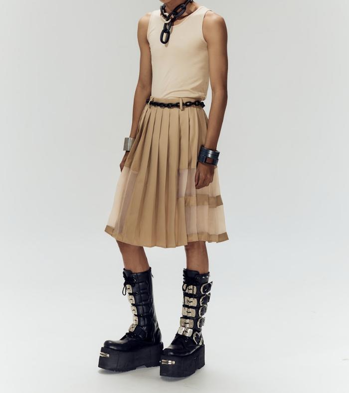 Person wearing a beige sleeveless top and pleated skirt with black platform boots on a white background