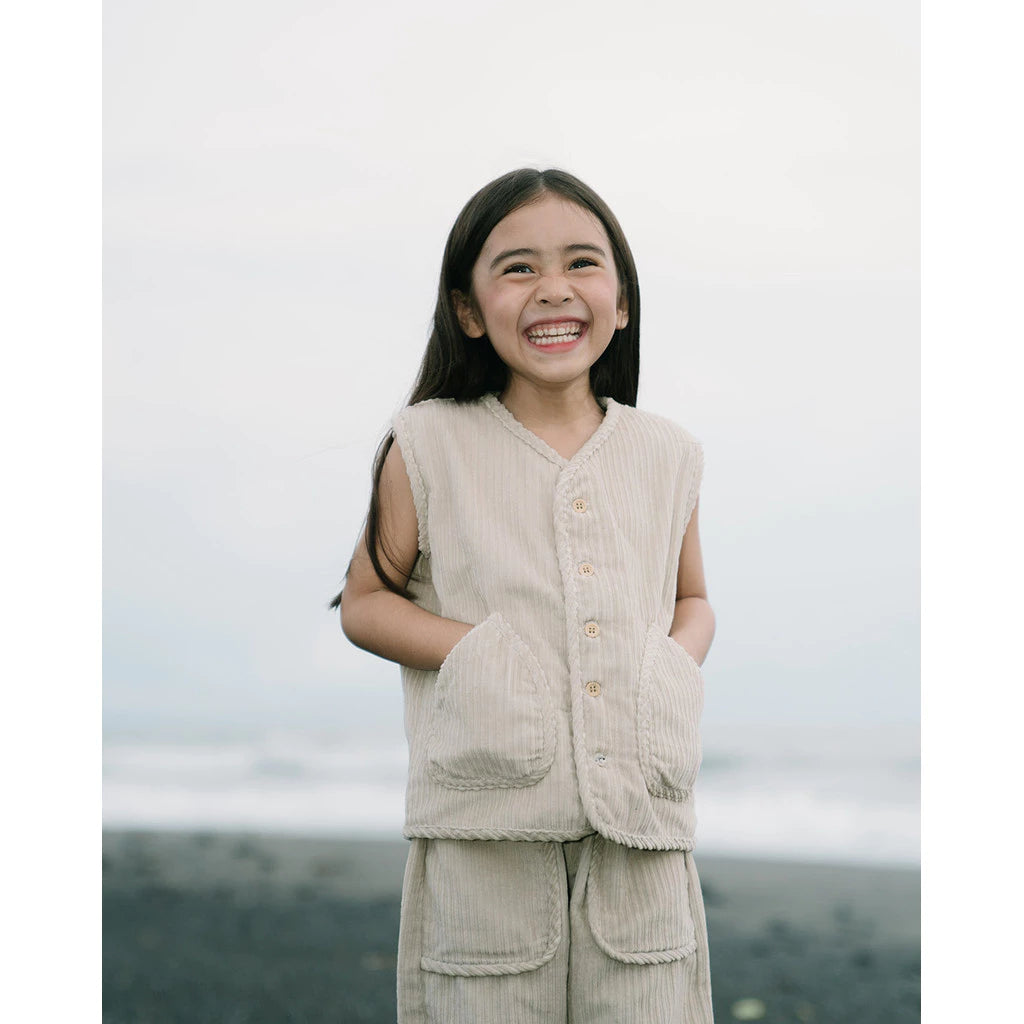 Young girl wearing a beige corduroy vest and pants standing against a blurred natural background