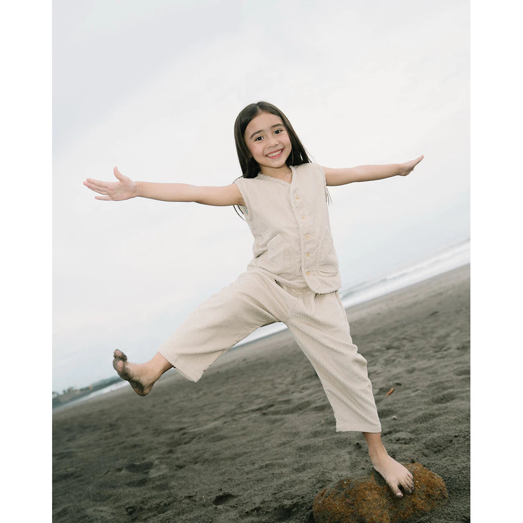 Child in beige vest and pants standing on a beach with arms outstretched