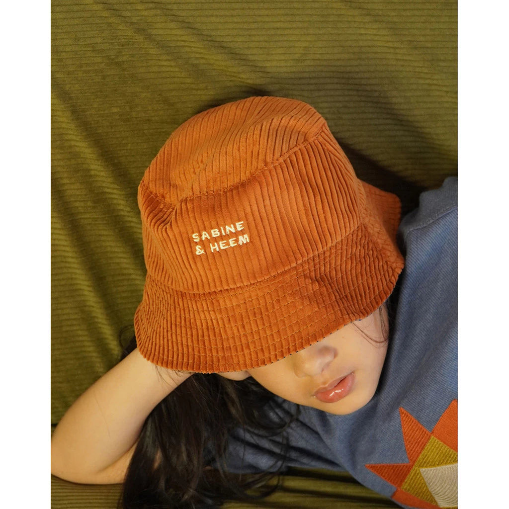 Kids wearing an orange bucket hat with 'Sabine & Heem' branding, lying on a green surface.