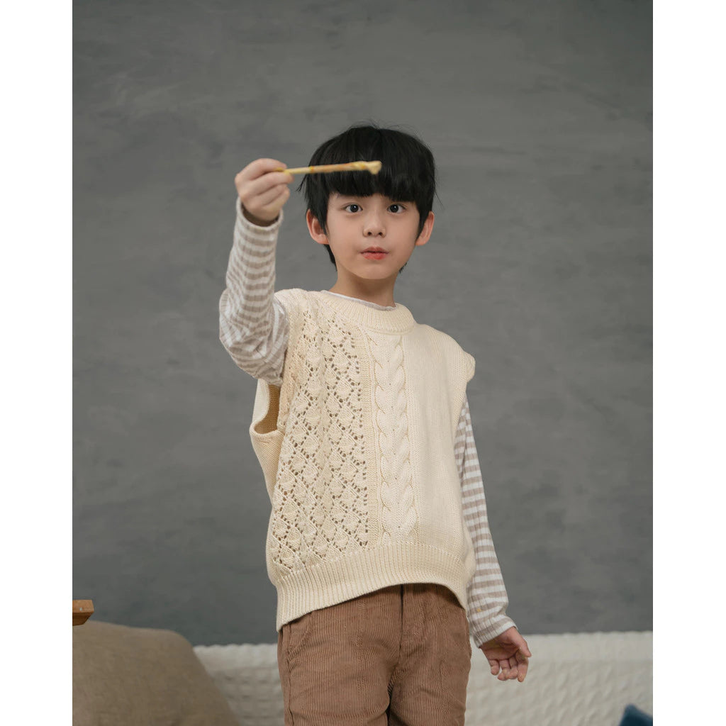 Child wearing a cream knitted vest and holding a wooden spoon against a gray background