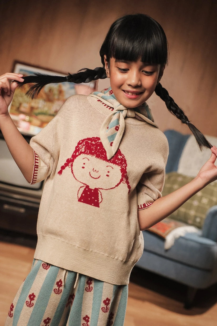 Young girl wearing a knitted tee with a red character design by Sabine & Heem kids clothing, standing in a room with a couch and framed picture.