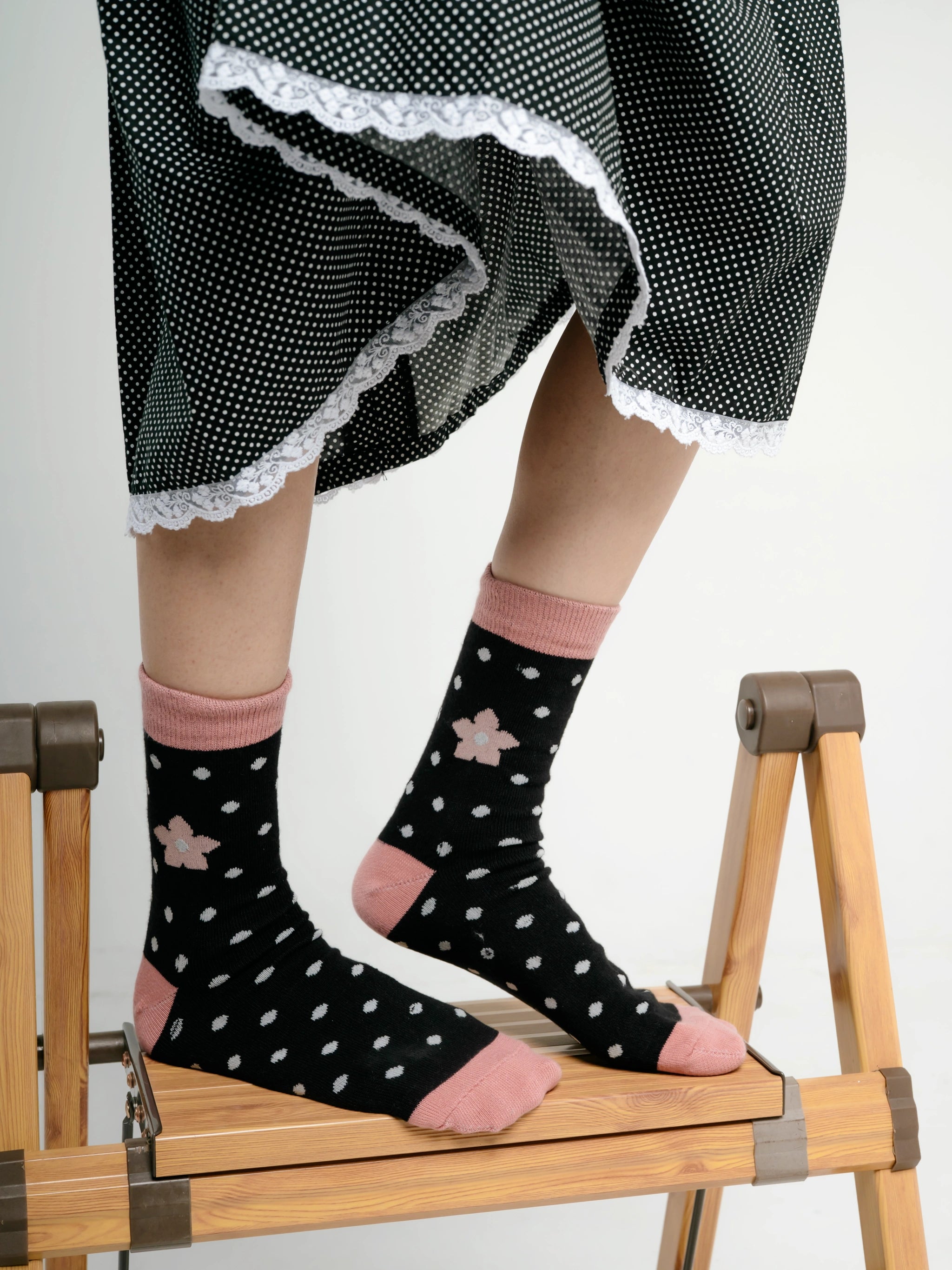 Person wearing black socks by Tome Ame with white polka dots and pink accents, sitting on a wooden chair.