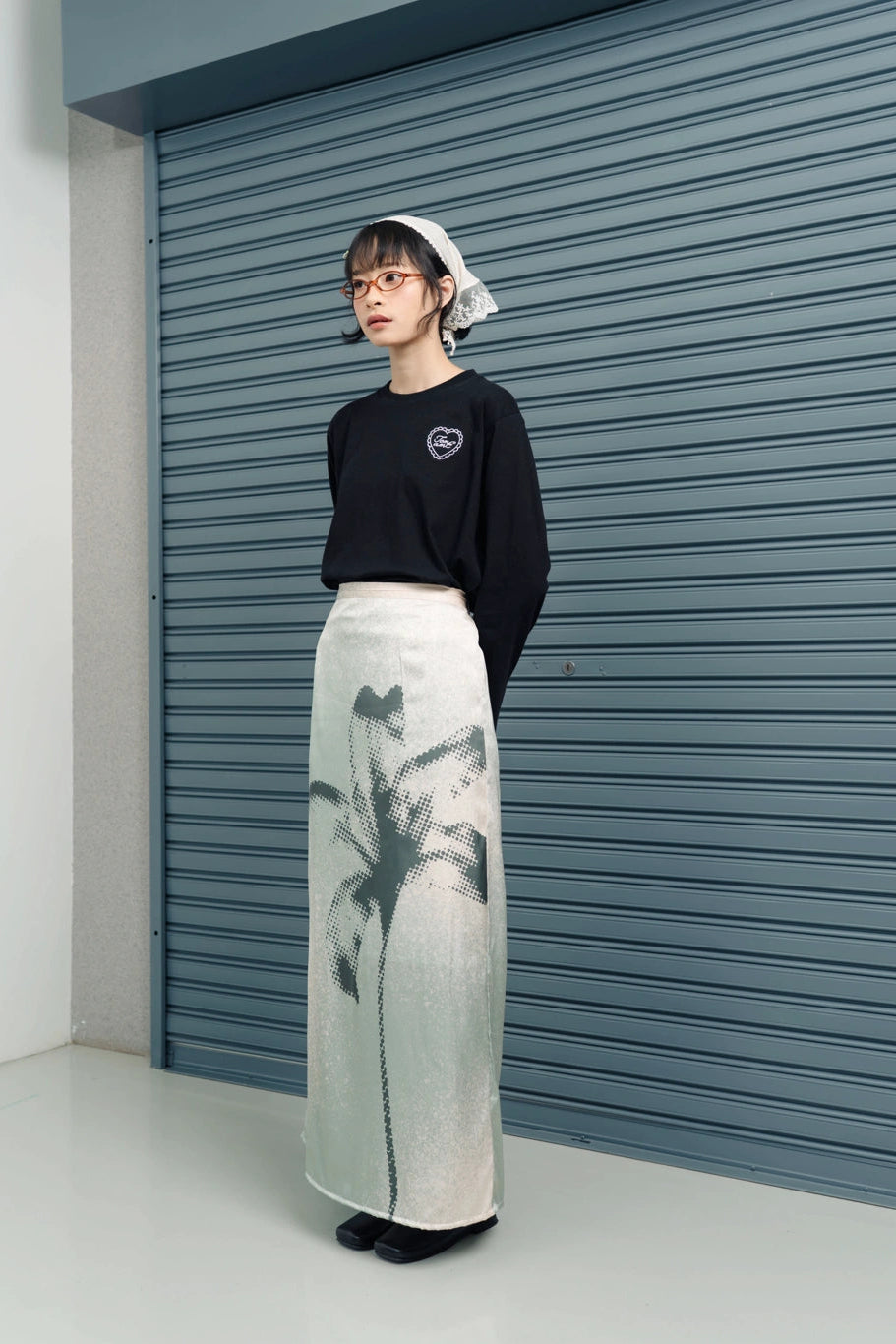 Person wearing a black top and ligh green skirt with a floral pattern by Tome Ame, standing in front of a blue shutter.