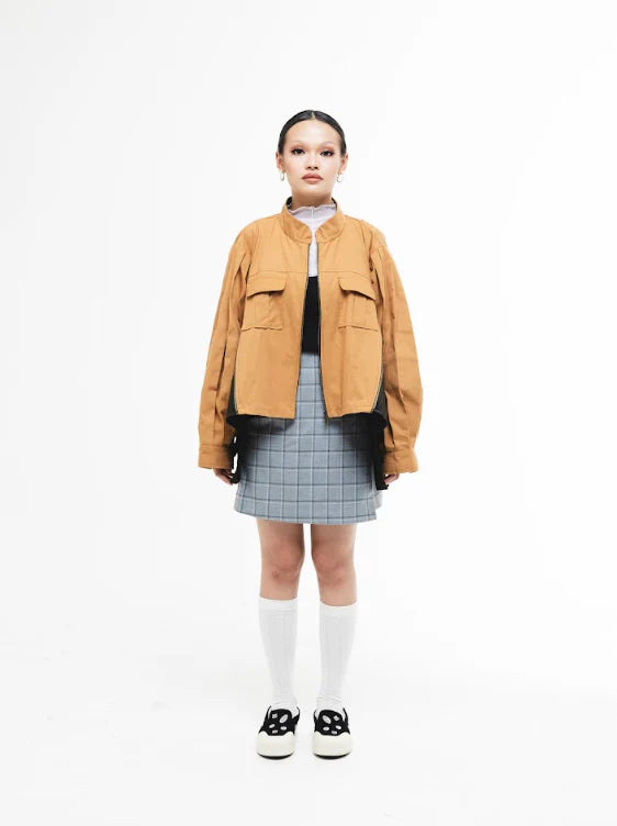 Person wearing a mustard yellow jacket by Wad Studio over a plaid skirt and white top on a white background.
