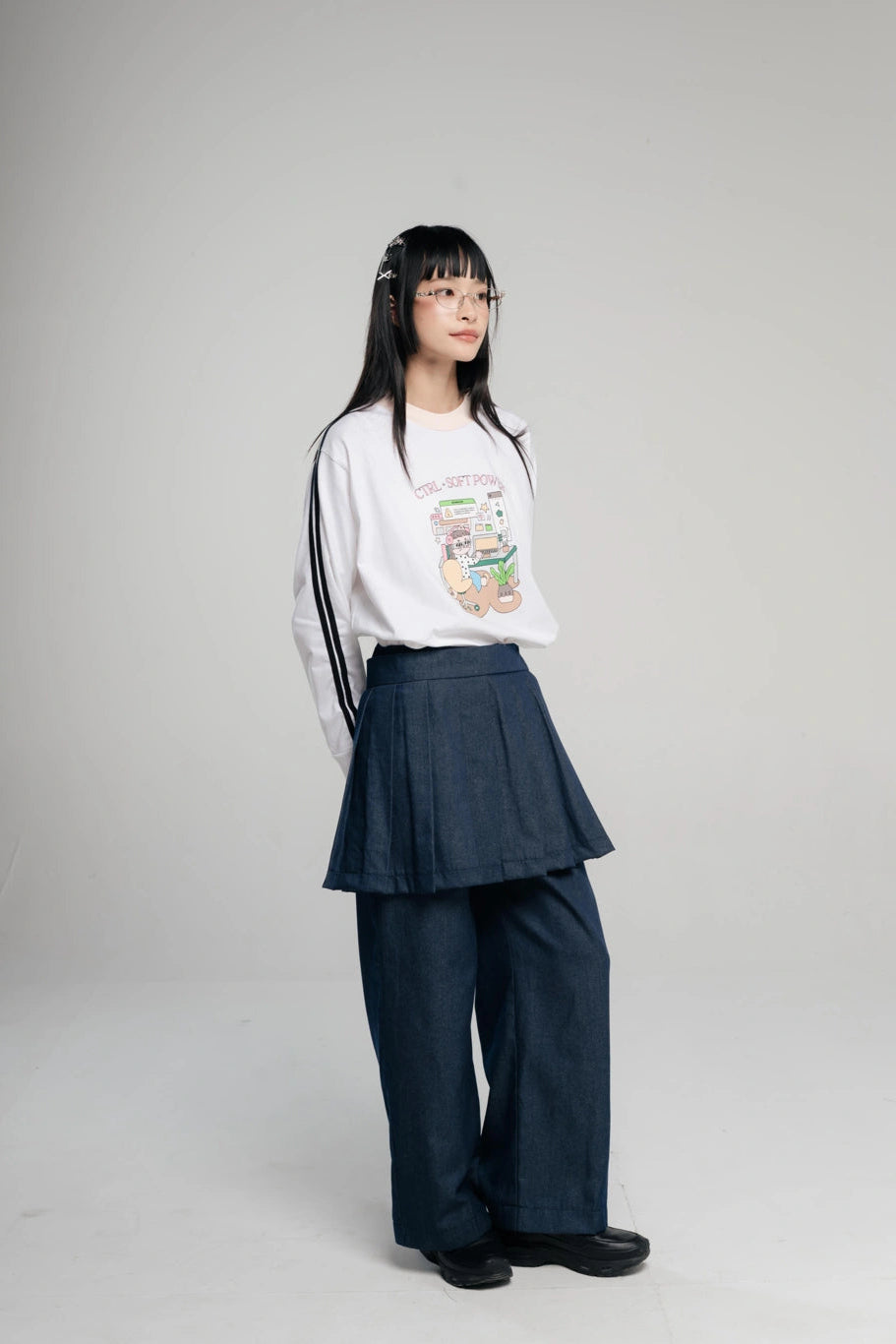 A young woman with long black hair and glasses stands against a light gray studio background, wearing a white long-sleeve graphic sweatshirt with black stripes on the sleeves, and a dark blue pleated layered pants by Tome Ame.