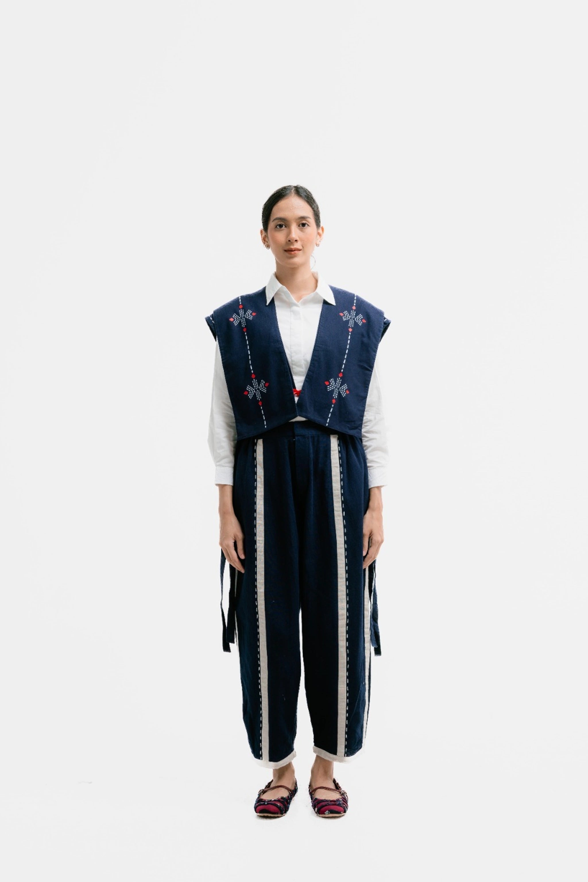 A woman wearing a navy and cream colored linen crop vest by Damakara with geometric patterns, over a white top, paired with dark pants and flat shoes.