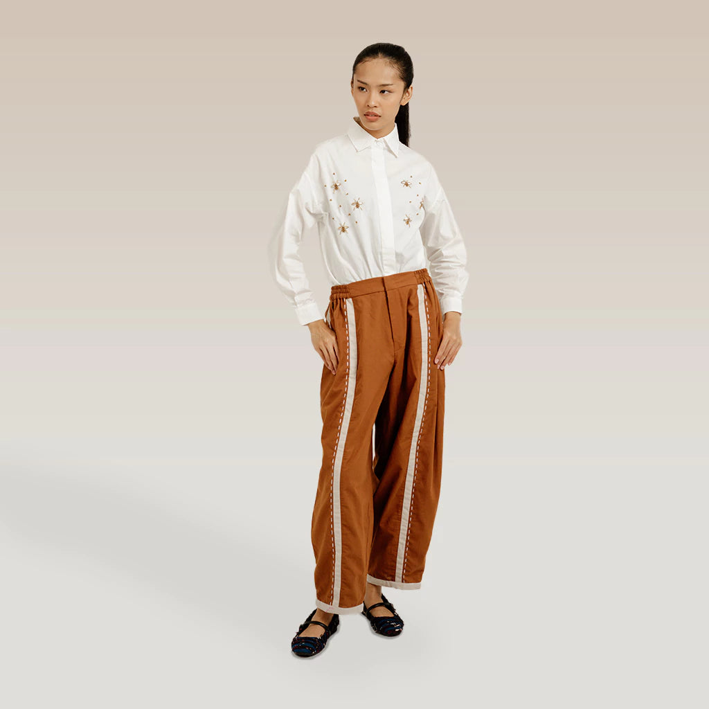 Person wearing a white blouse and brown pants by Damakara with white trim on a plain background
