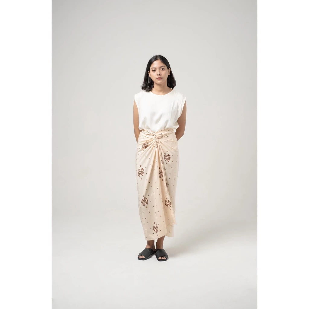 A woman standing wearing a traditional broken white skirt with a brown pattern called Kaimana Skirt from Damakara, paired with a white top.