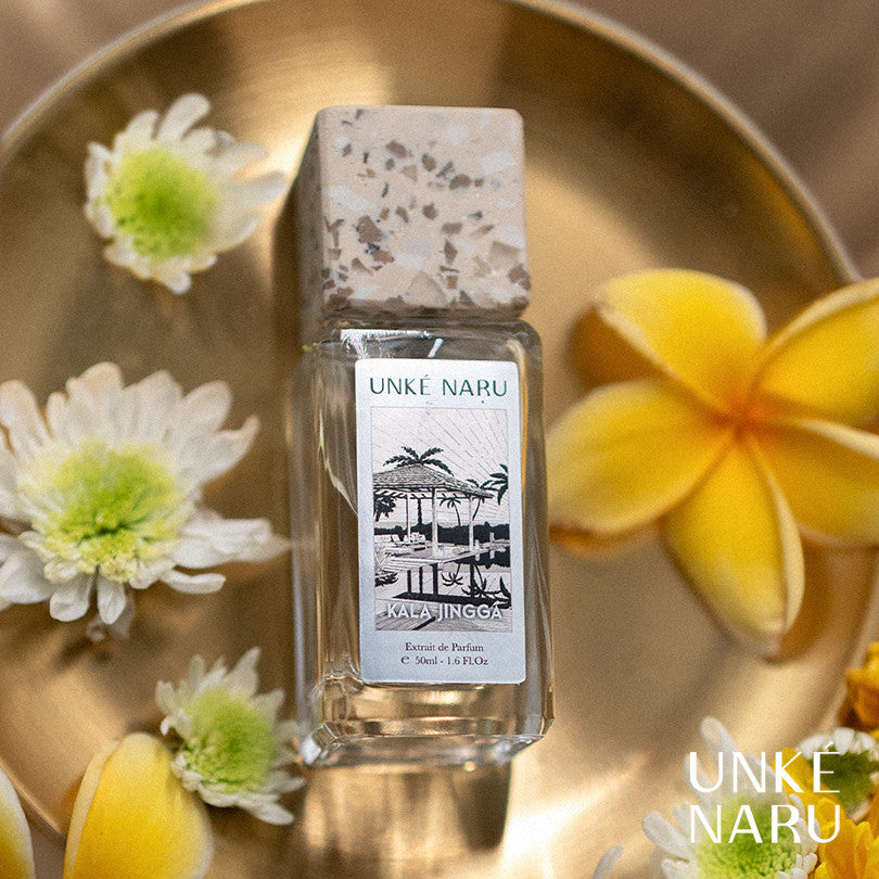 A bottle of Kala Jingga perfume by Unke Naru, placed on a surface with flowers around it, notably a yellow frangipani flower.