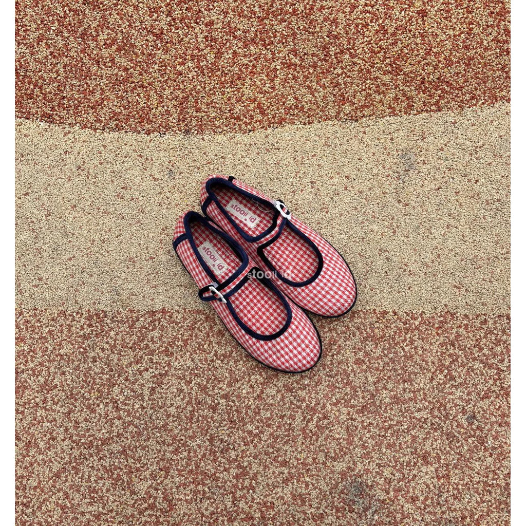Pair of red and white checkered flat shoes with black straps by Stooli on a multicolored textured surface