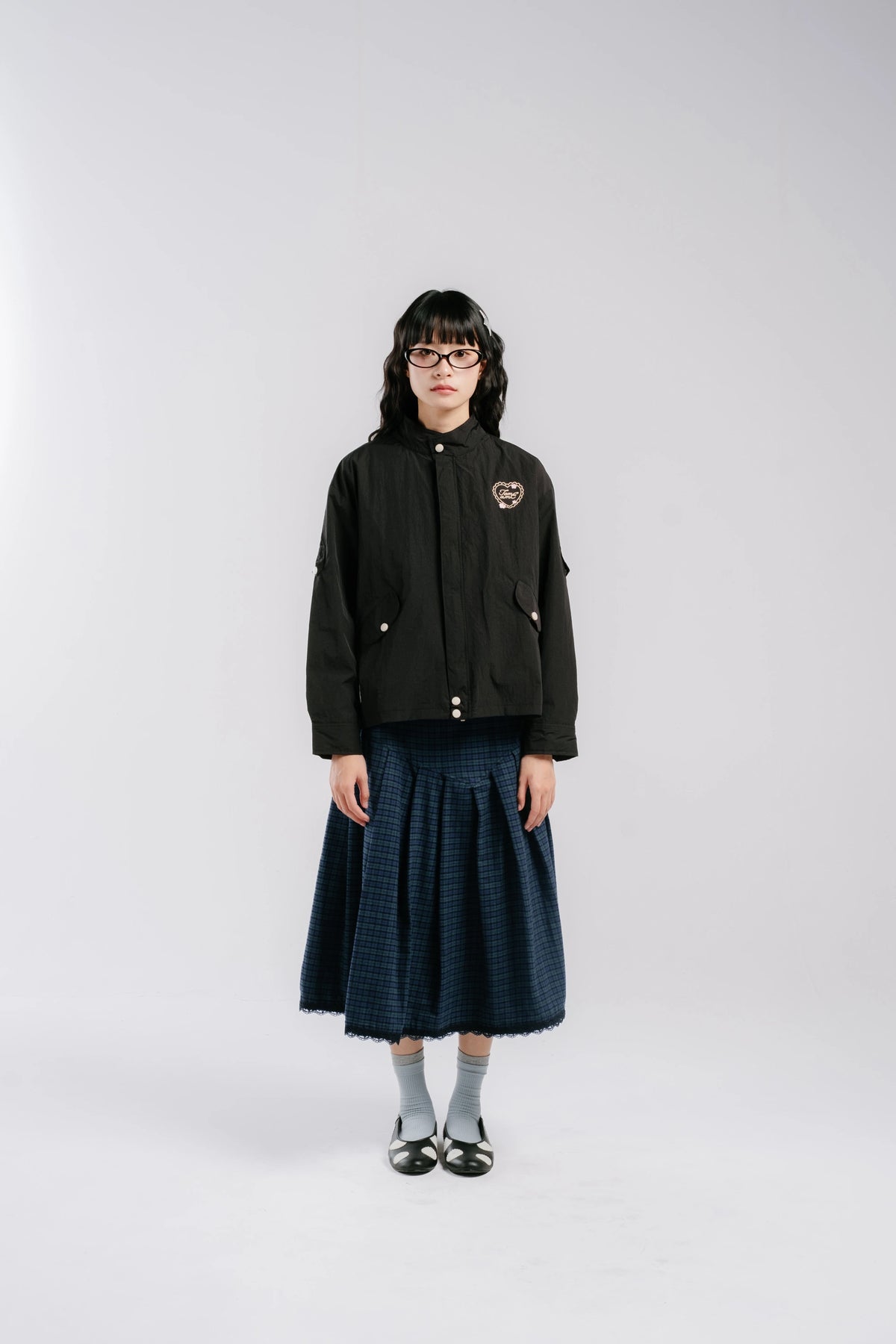 A model stands against a light gray studio background wearing a black jacket by Tome Ame with a small embroidered chest patch, a dark blue pleated skirt, gray socks, and black shoes.