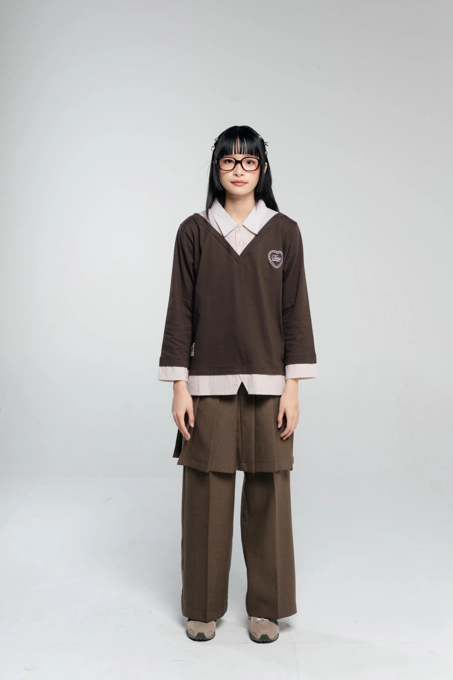 A model wearing a long-sleeve v-neck layered shirt by Tome Ame, matching brown pleated skirt, and wide-leg trousers, standing against a light gray background.