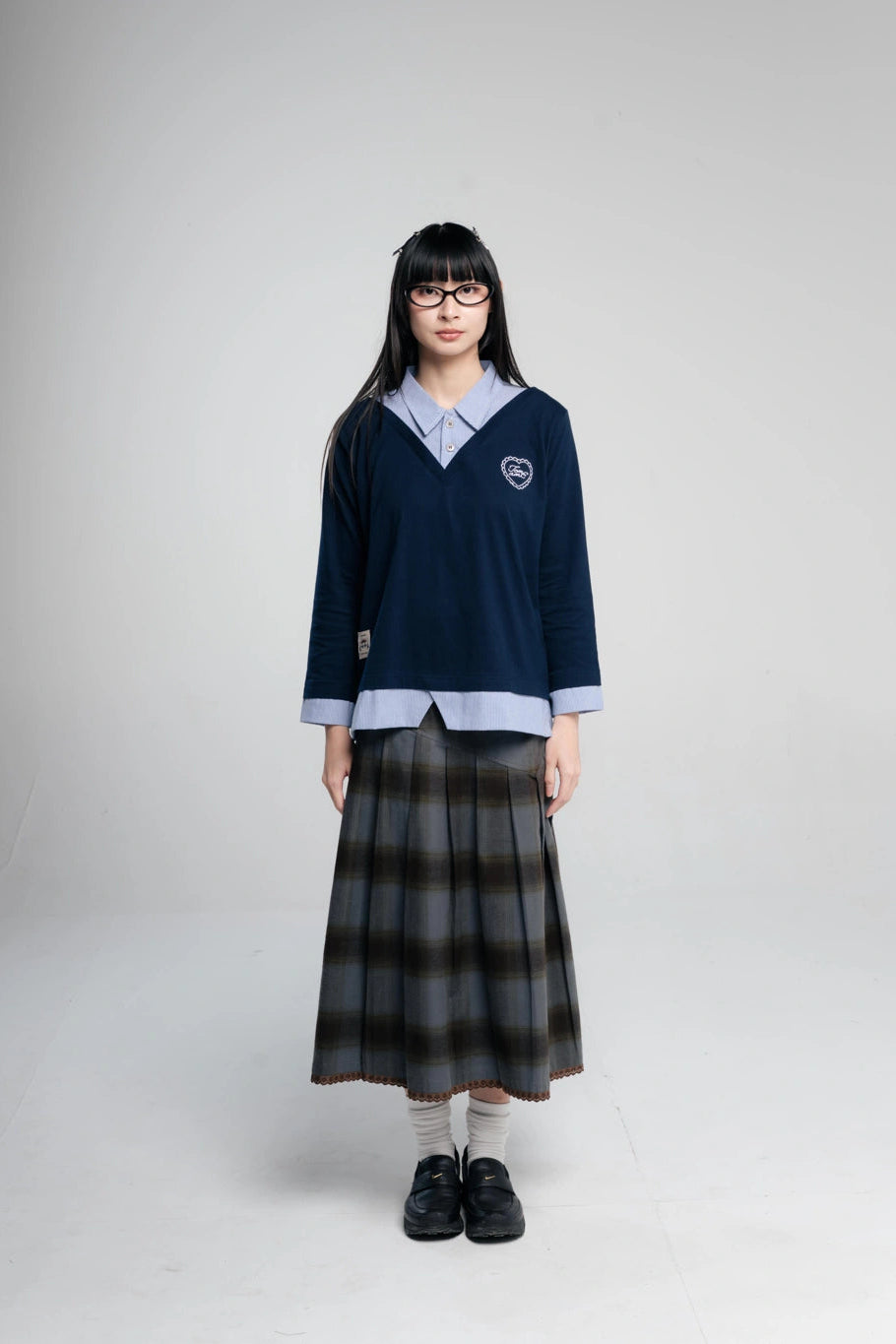 A young woman modeling a navy blue layered shirt by Tome Ame, paired with a dark gray and black plaid pleated skirt, white socks, and black loafers, against a plain light gray studio background.