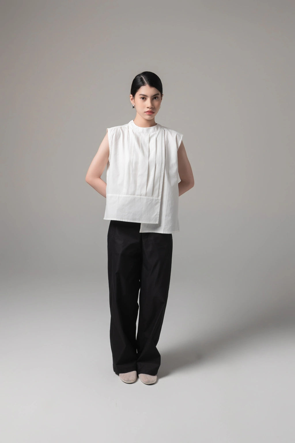 Woman wearing a broken white sleeveless blouse by Kajuaruna and black pants on a gray background